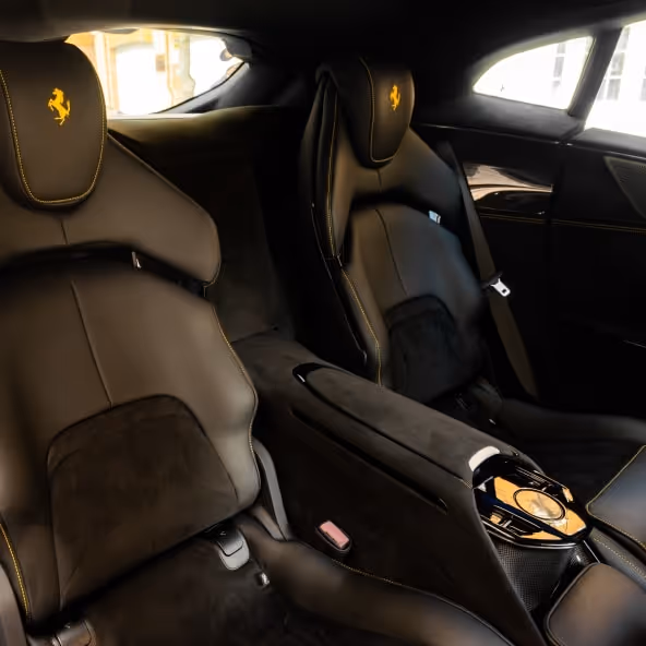 A view of the individual luxury rear seats in the Ferrari Purosangue, featuring black Alcantara, yellow stitching, and the Ferrari logo embroidered on the headrests.