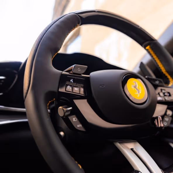 A close-up of the Ferrari Purosangue steering wheel featuring integrated driving controls, paddle shifters, and the yellow Ferrari shield.