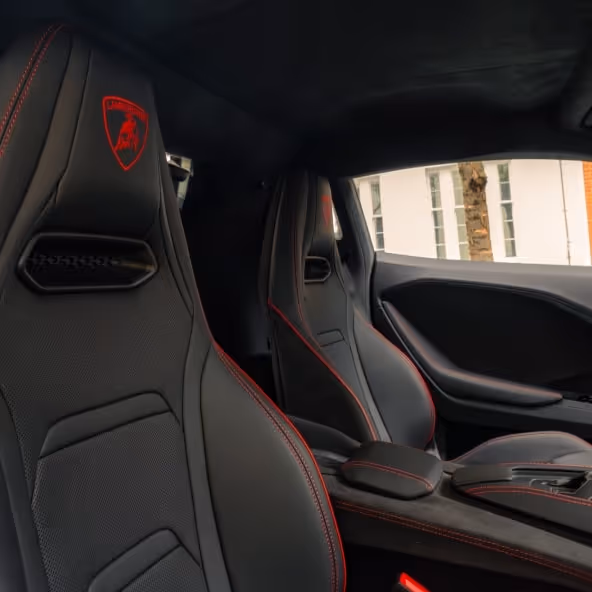 A close-up of the black leather and Alcantara racing seats inside a Lamborghini, featuring red contrast stitching and the embroidered red bull emblem