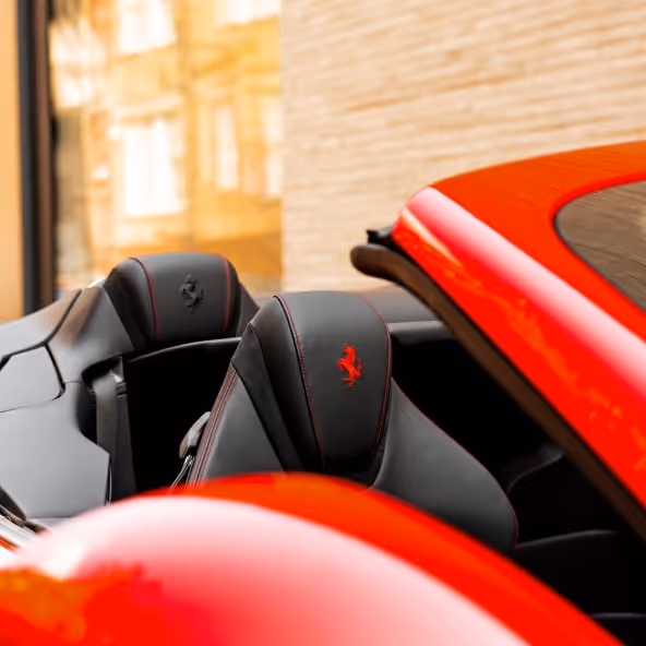 A close-up of the Ferrari Roma Spider interior showing black leather sport seats with embroidered Ferrari prancing horse logos and premium red stitching details.