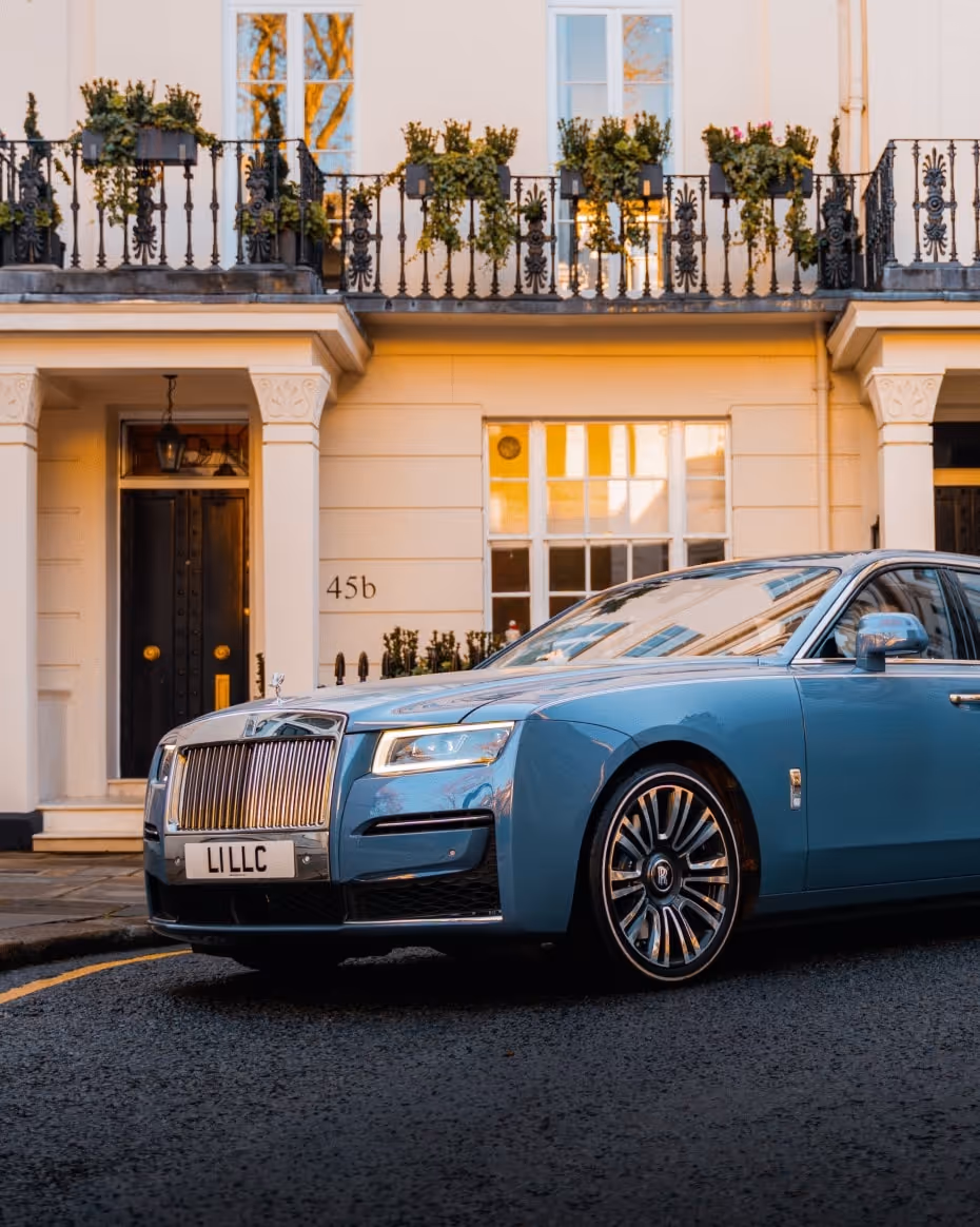 The Rolls-Royce Ghost in bespoke light blue/grey, parked elegantly outside a classic white stucco London residence, emphasizing LLC's discreet luxury chauffeur service.