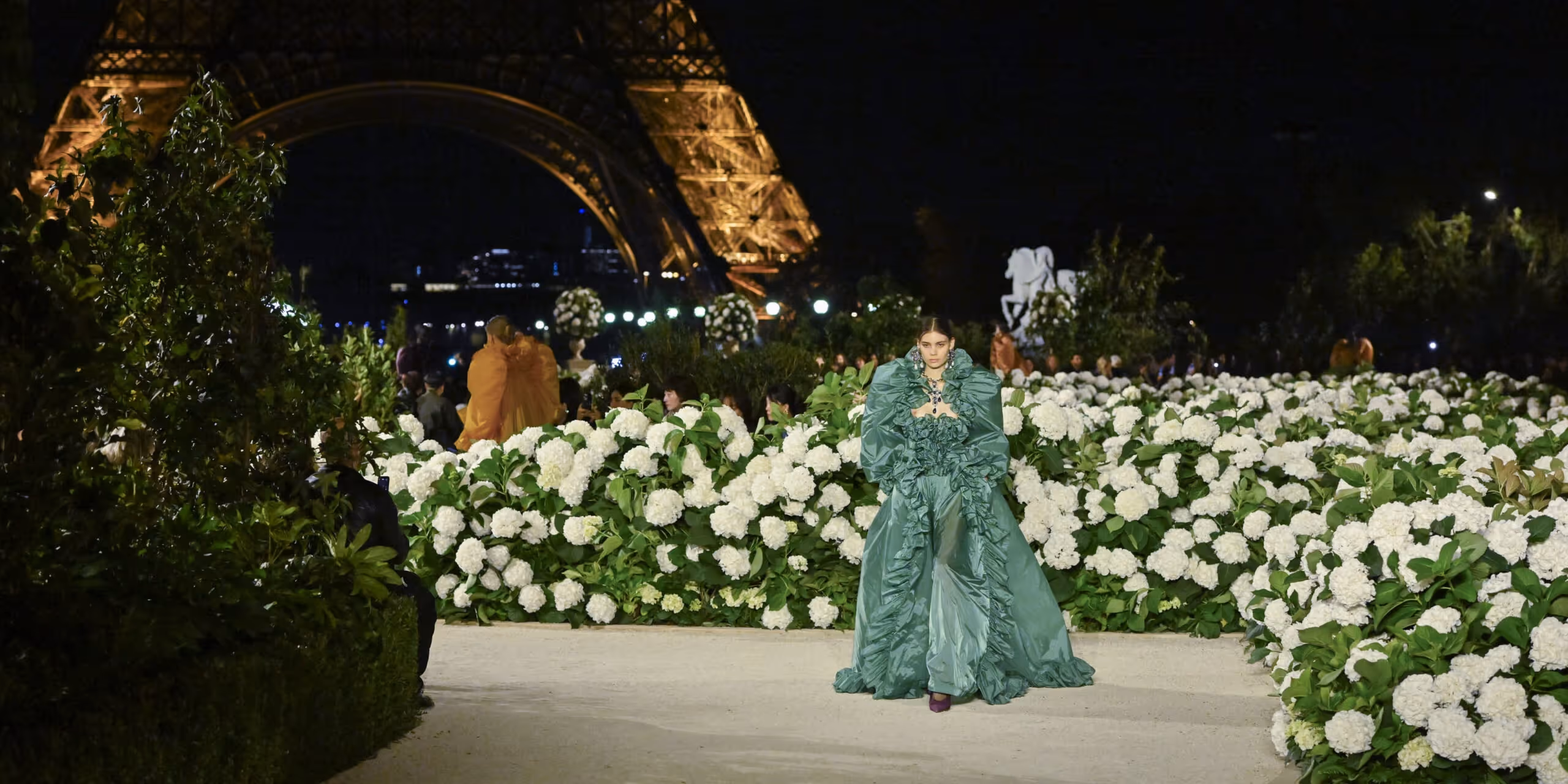 Model walking through a white floral runway installation beneath the illuminated Eiffel Tower during Paris Fashion Week at night.