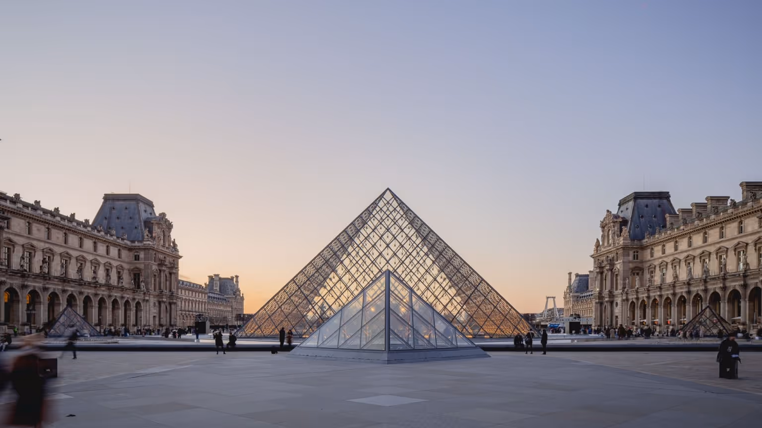 The Louvre Pyramid in Paris at sunset with the historic museum architecture surrounding the glass structure.