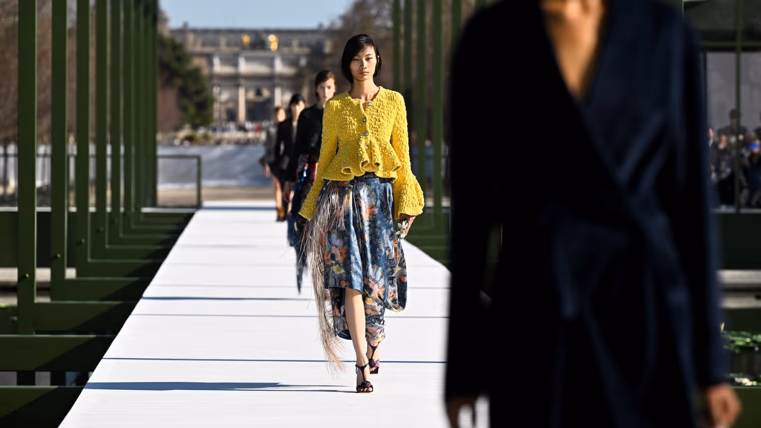 Models walking down an outdoor runway during Paris Fashion Week with modern architectural framing and luxury fashion styling.