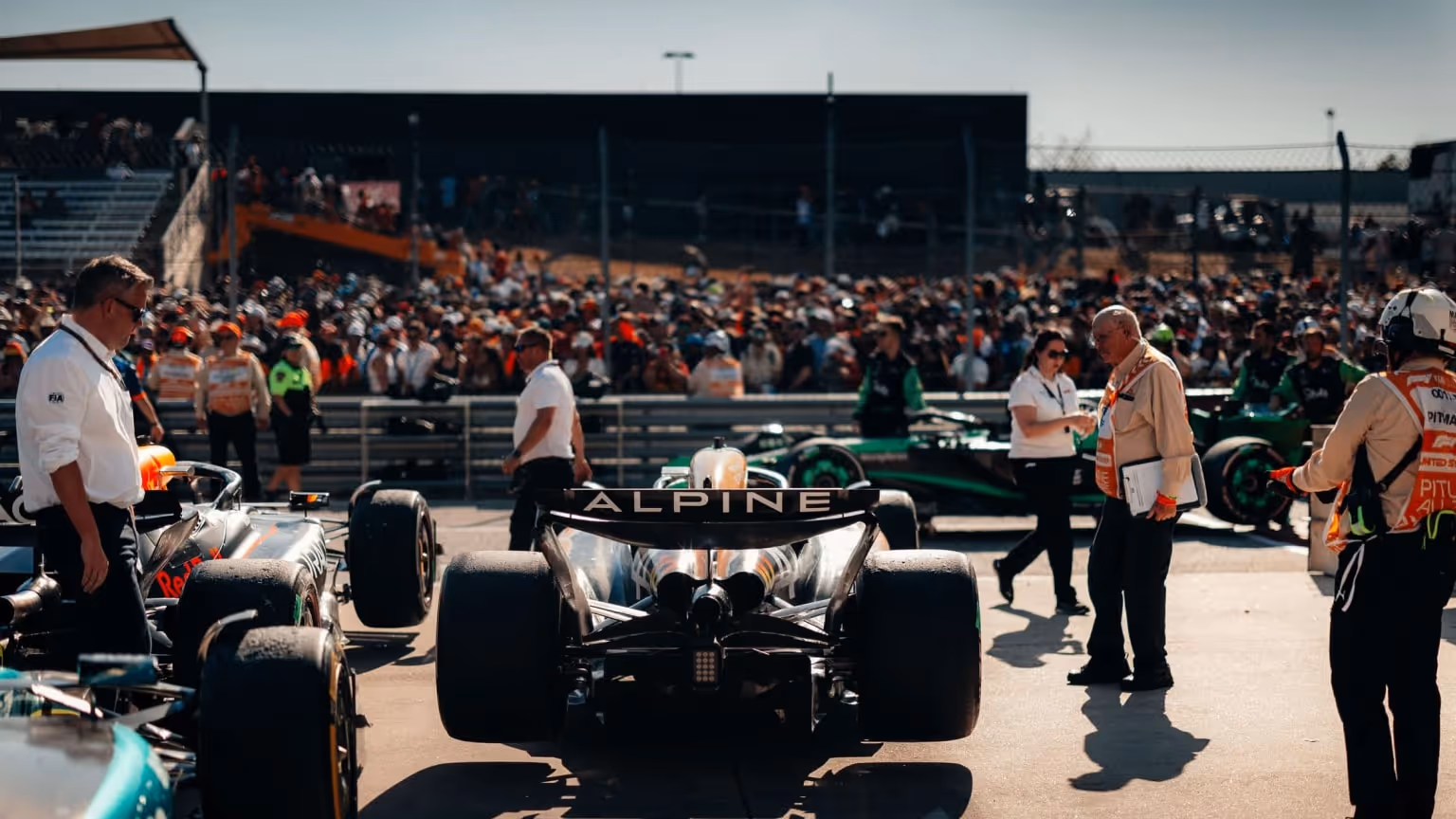 Formula 1 cars lined up on the starting grid before a race, with Alpine, Red Bull Racing, and Aston Martin cars visible. Team personnel and FIA officials surround the cars; spectator grandstands fill the background.