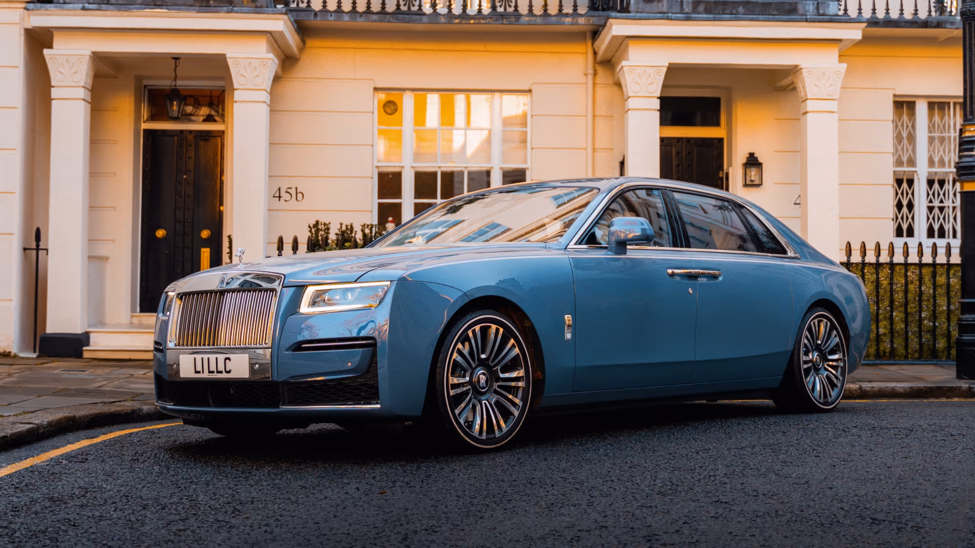 The Rolls-Royce Ghost in bespoke light blue/grey, parked elegantly outside a classic white stucco London residence, emphasizing LLC's discreet luxury chauffeur service.