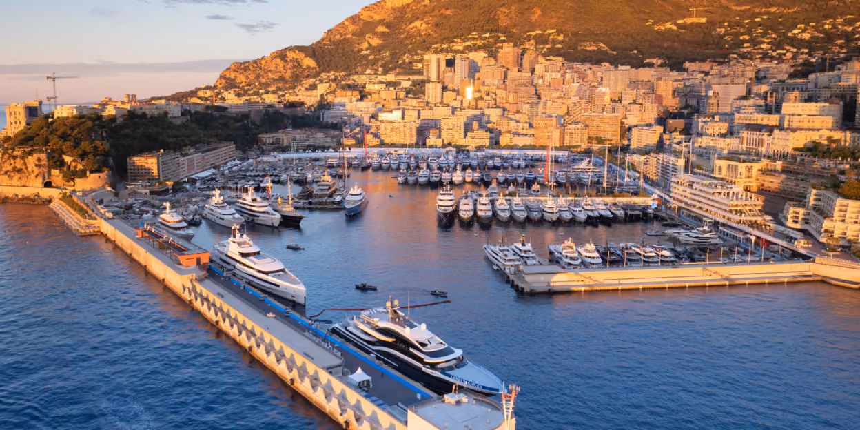 Superyachts moored along Quai Rainier III at Port Hercule in Monaco during the Monaco Yacht Show, with the Principality's coastline visible in the background.