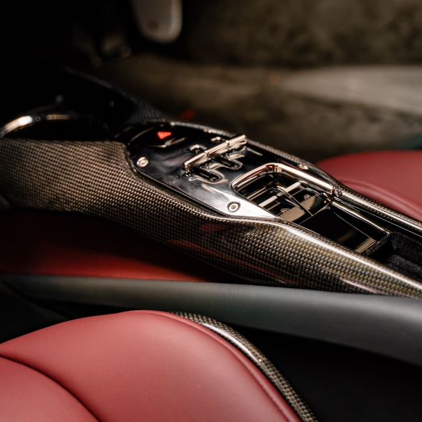 Close-up of the Ferrari 296GTB carbon-fibre centre console with metallic controls, gloss-black trim, and Rosso leather seats.