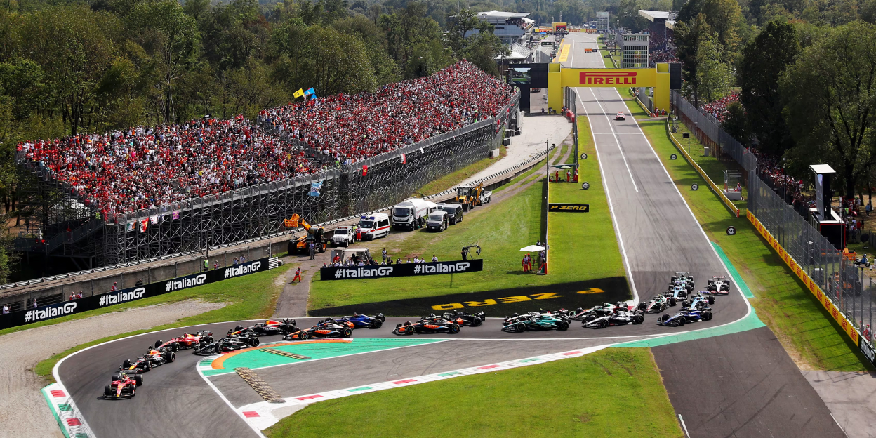 Aerial view of Formula 1 cars racing through the opening corner at Monza during the Italian Grand Prix, with packed grandstands and the main straight in the background.