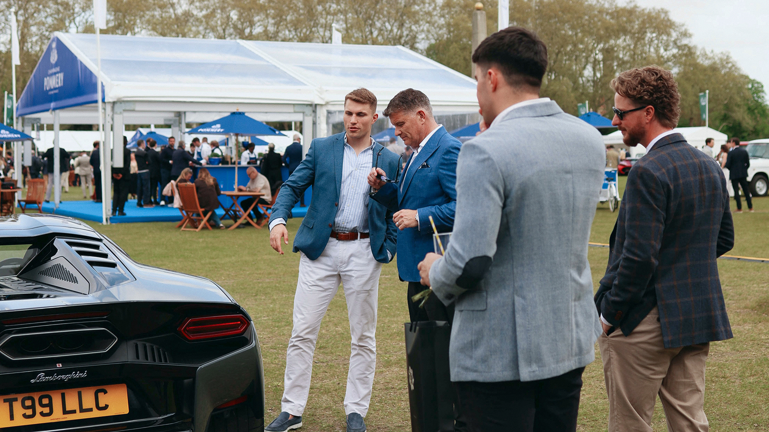 A group of four men standing and talking beside a black Lamborghini Temerario on a grass lawn at an outdoor luxury automotive event, with hospitality tents and guests in the background.