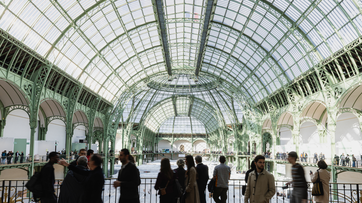 Interior of the Grand Palais in Paris with its monumental glass-and-iron roof, ornate green metalwork and exhibition stands filled with visitors.