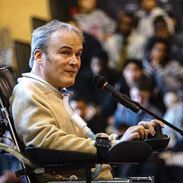 Man in wheelchair speaking into microphone before an audience in a blurred background.