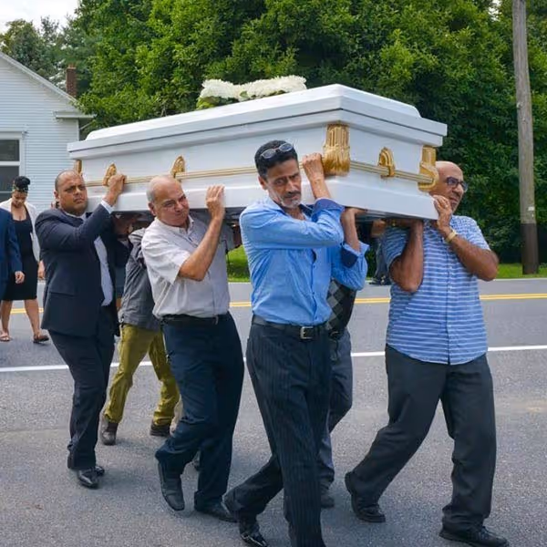 Six men carrying a white coffin with gold accents on a road, surrounded by trees and houses.