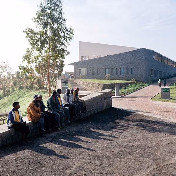 Group of people sitting on a stone wall casting long shadows near modern buildings on a sunny day.