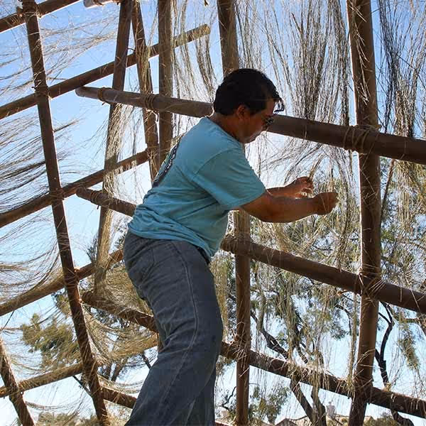 Man in blue shirt weaving or repairing a large natural fiber net or wall on a wooden frame outdoors.