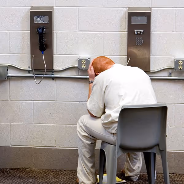 Person in white clothes sitting on a chair with head in hands near two metal phone booths on a wall.