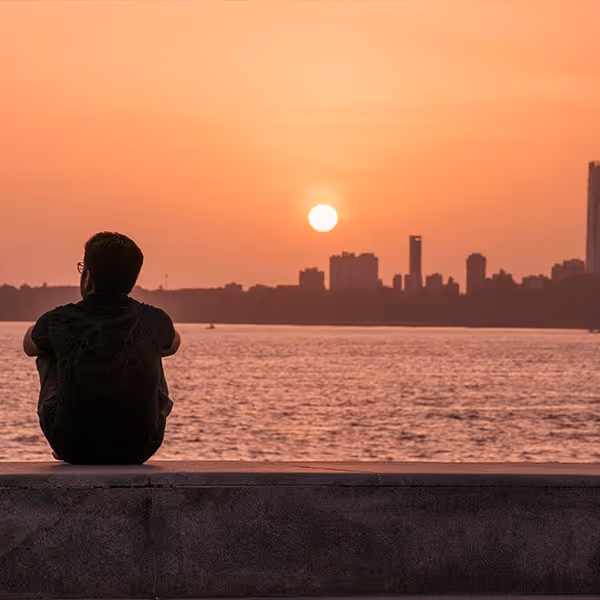 Person sitting on a ledge looking at the sunset over water with a city skyline in the background.