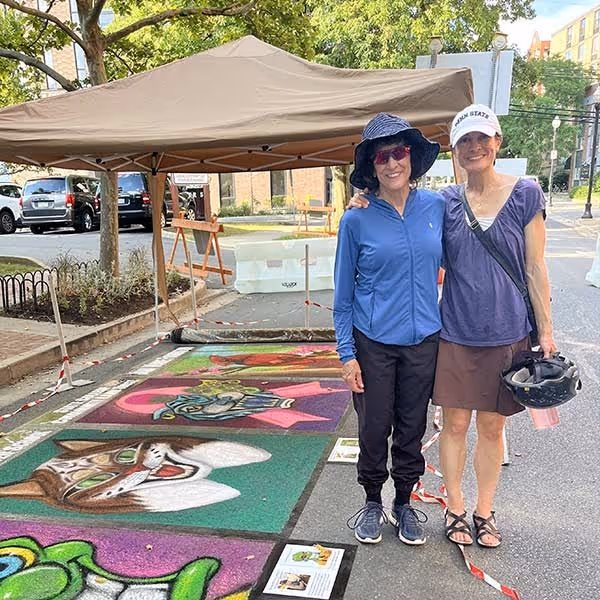 Two women standing on sidewalk with colorful chalk art of animals under a canopy tent.