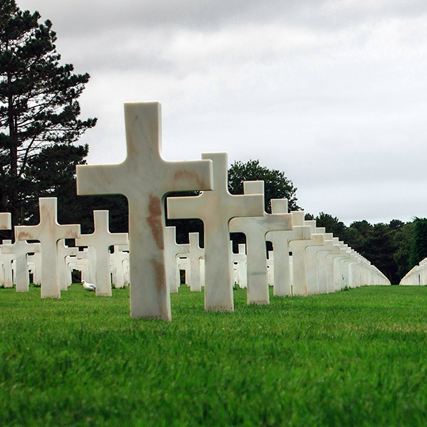Rows of white marble crosses in a cemetery on green grass with trees and a cloudy sky.