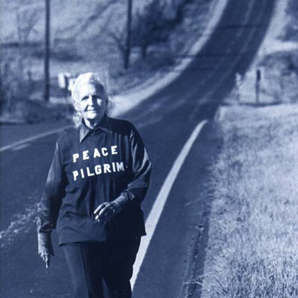 Older woman walking alone on a road wearing a shirt that says Peace Pilgrim.