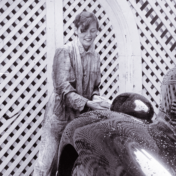 Person wearing a smock polishing a shiny, curved surface in front of a lattice backdrop.