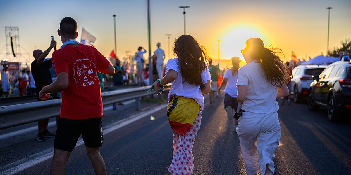 People running on a road at sunset during an outdoor event with cars and spectators nearby.