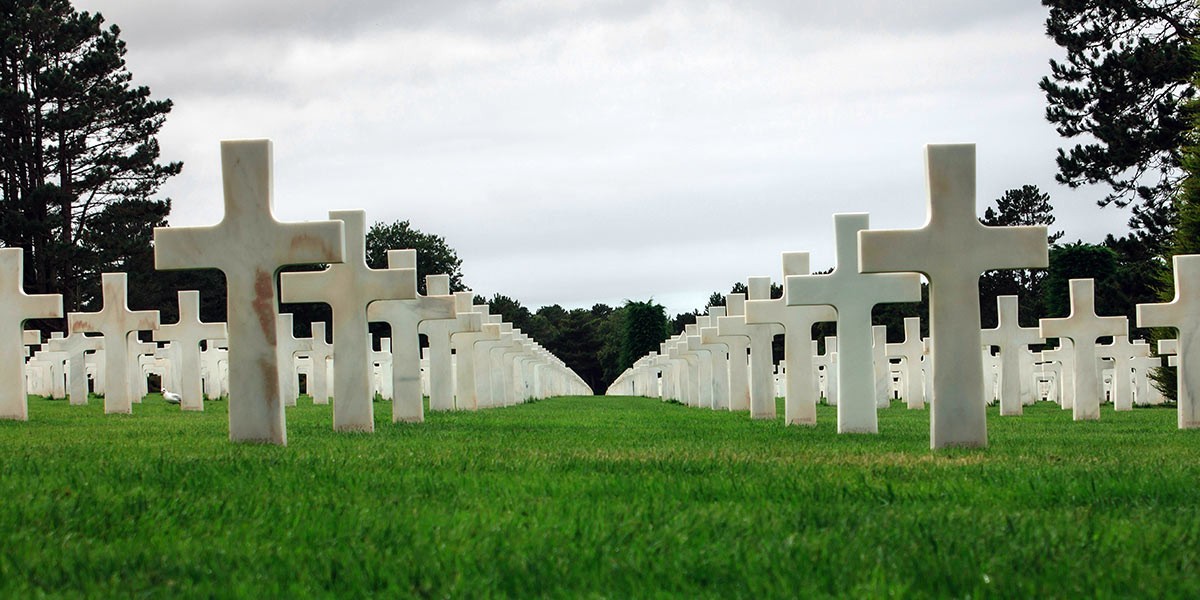 Rows of white crosses marking graves in a cemetery on green grass under a cloudy sky.