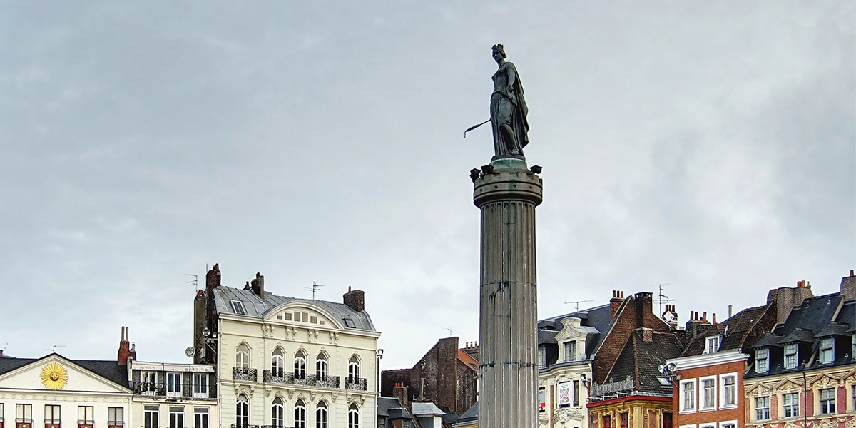 Statue of a crowned figure holding a staff atop a tall column with European buildings in the background.
