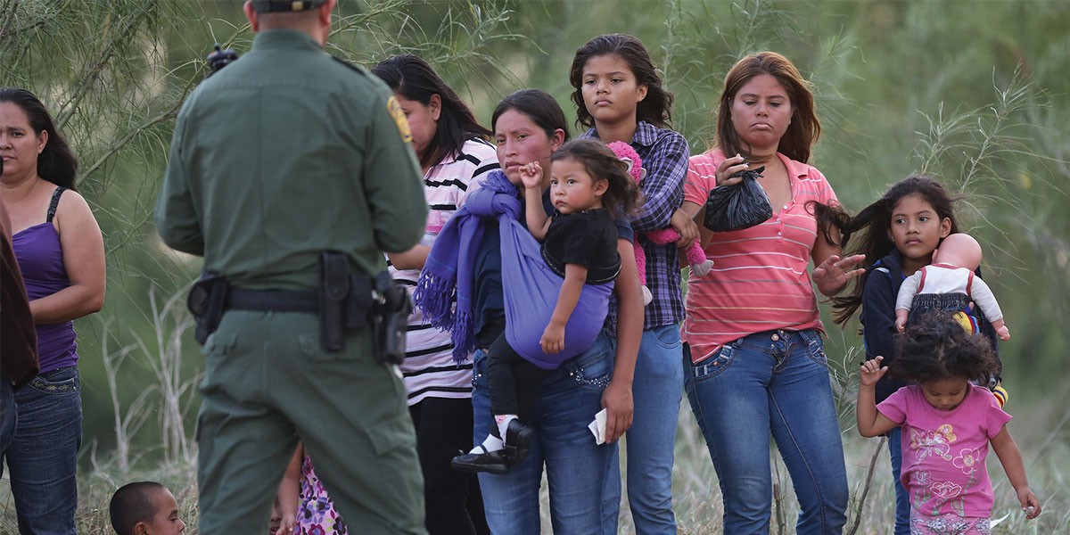 Group of women and children, some carrying infants, facing a uniformed officer in an outdoor setting.