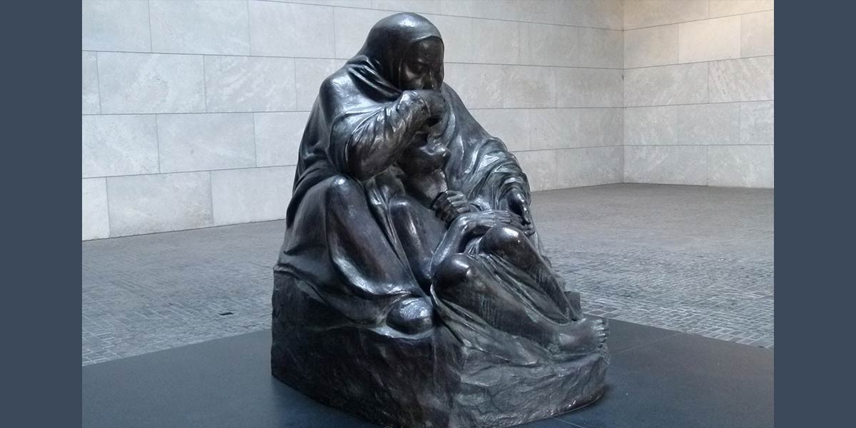 Dark bronze sculpture of a seated woman holding a limp, seated figure in her arms in a stone room.