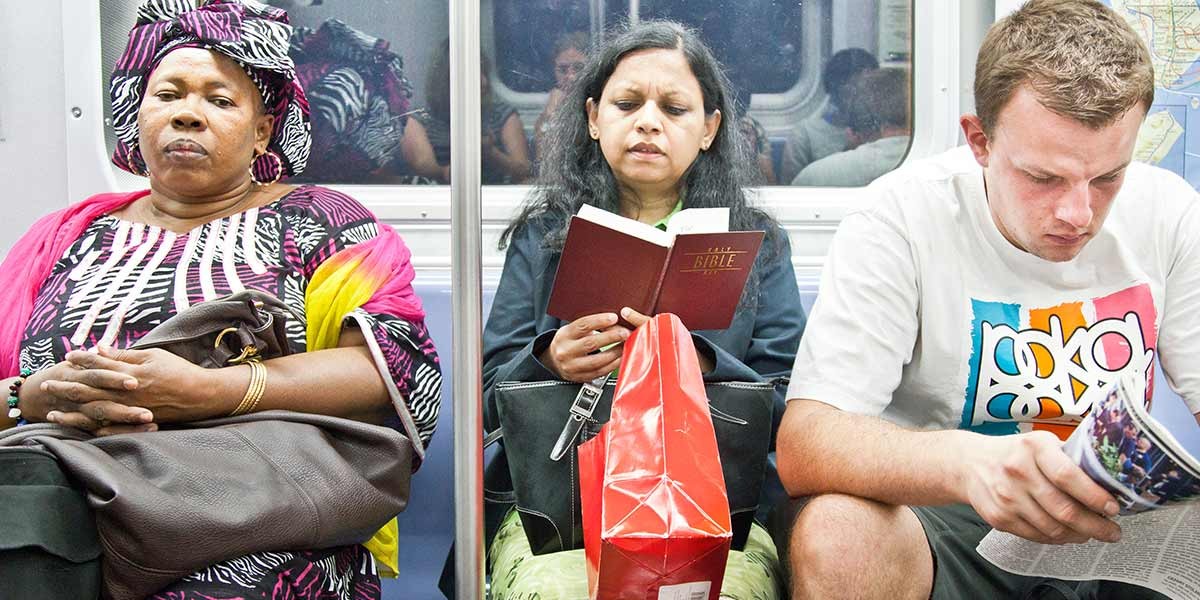 Three people sitting on a subway bench: woman in patterned clothes, woman reading Bible, man reading a magazine.