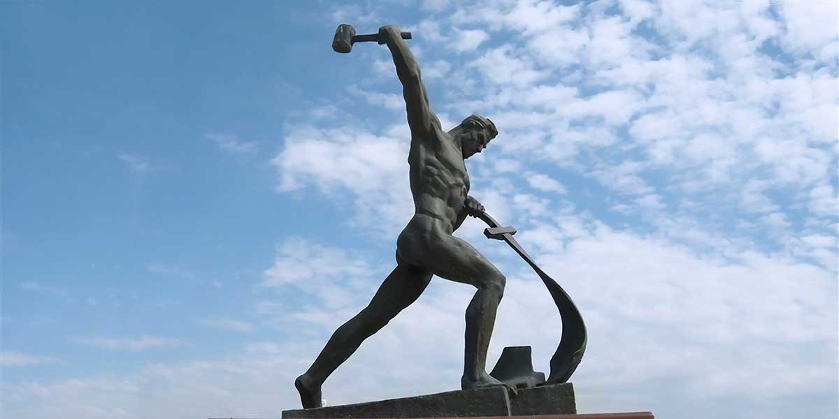 Bronze statue of a muscular man swinging a hammer with anvil against a partly cloudy sky.