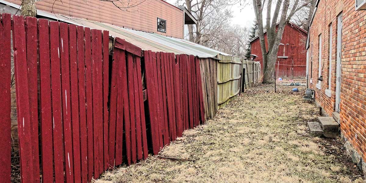 Leaning, weathered red wooden fence next to a brick building and dry grass in a backyard.