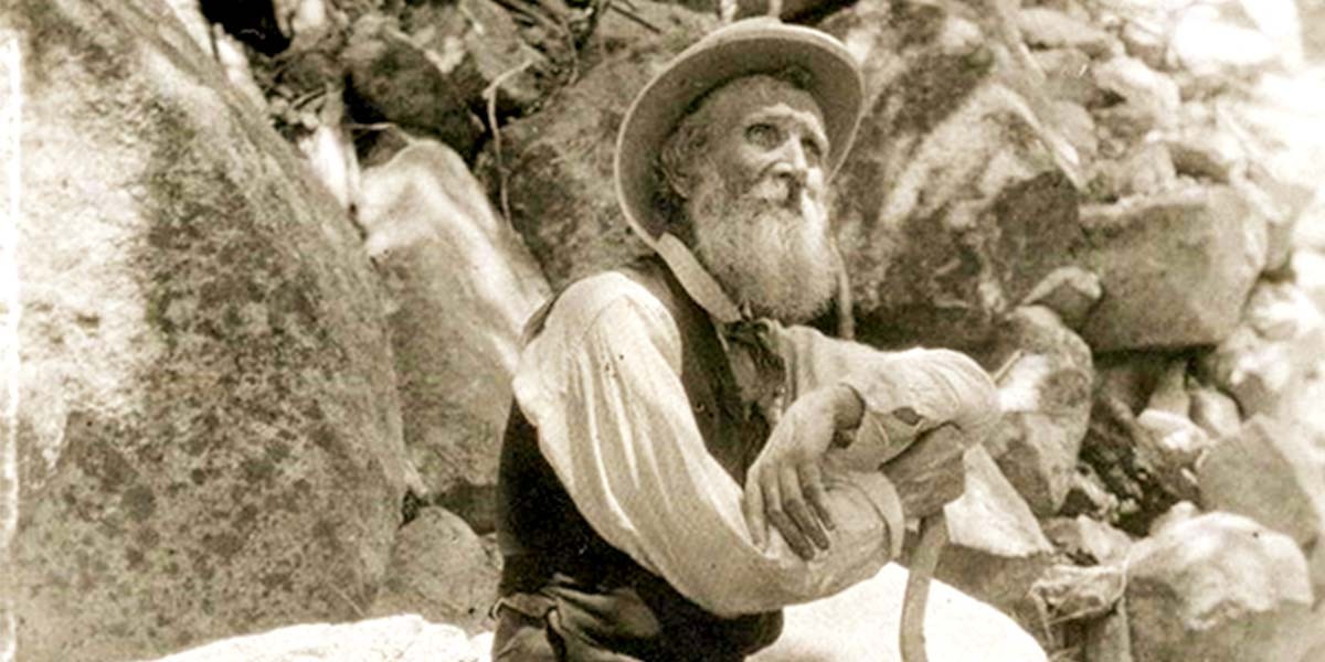 Bearded man in old-fashioned clothes and hat sitting on rock among large boulders.