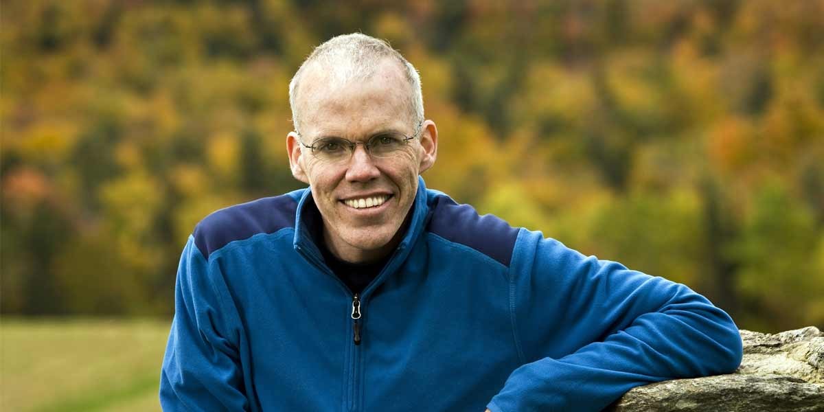 Smiling man with glasses in a blue jacket leaning on a stone with autumn foliage behind him.