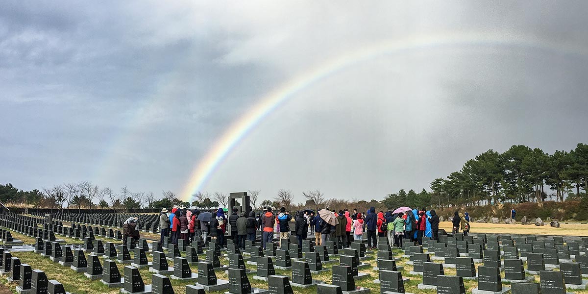 Group of people gathered at a cemetery with many gravestones under a cloudy sky and a rainbow.