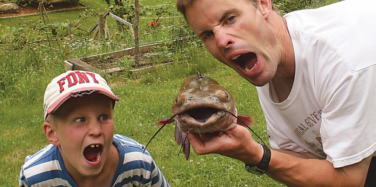 Man and boy outdoors holding a large fish, both with mouths wide open in playful expressions.
