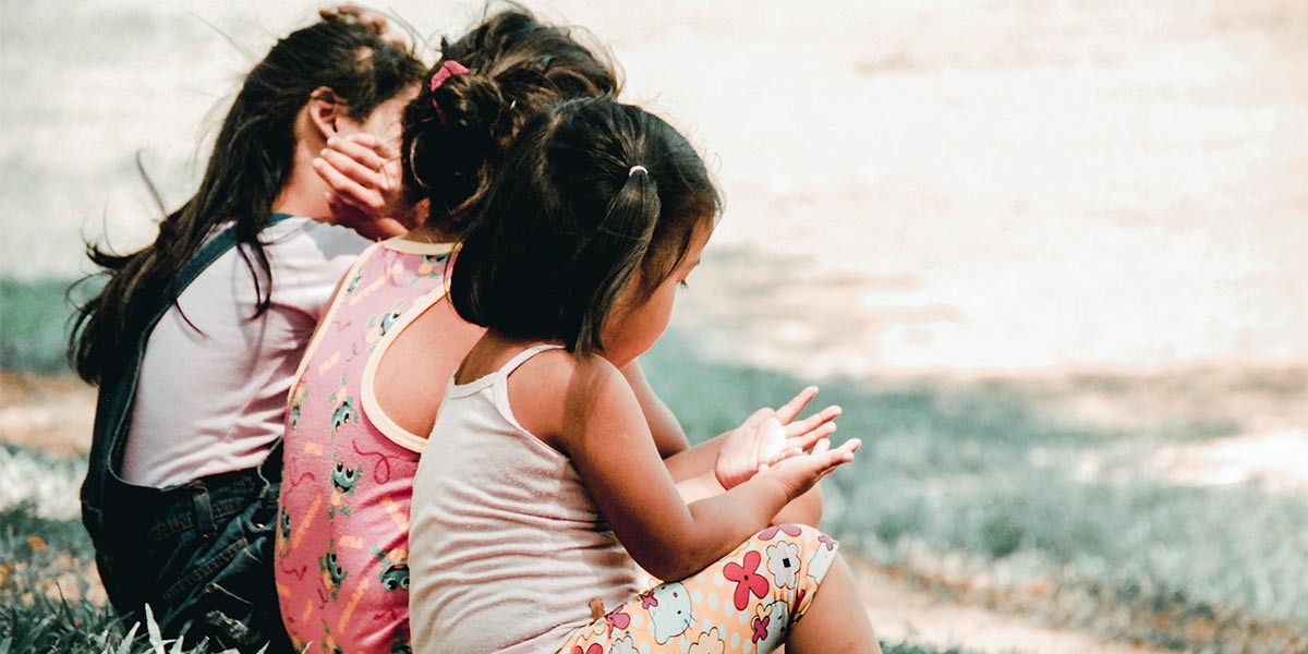 Three young girls sitting on grass outdoors, viewed from the side and back.