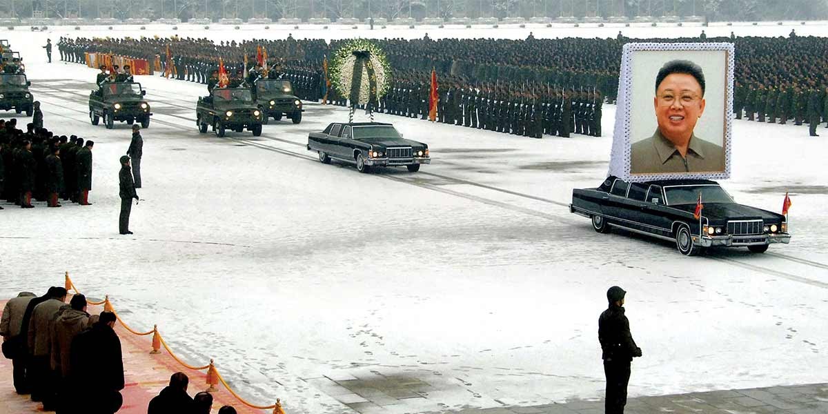 Military funeral procession with large portrait of a man, wreath, and soldiers in formation on snow-covered ground.