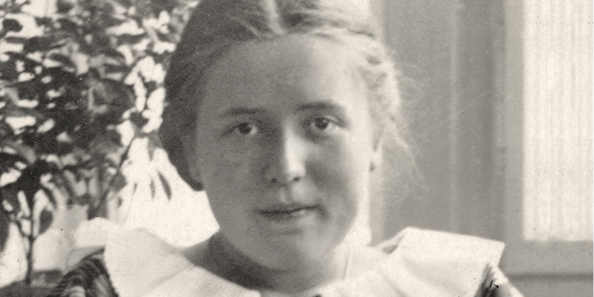 Close-up of a woman with light hair and a large white collar, seated indoors near a window and plant.
