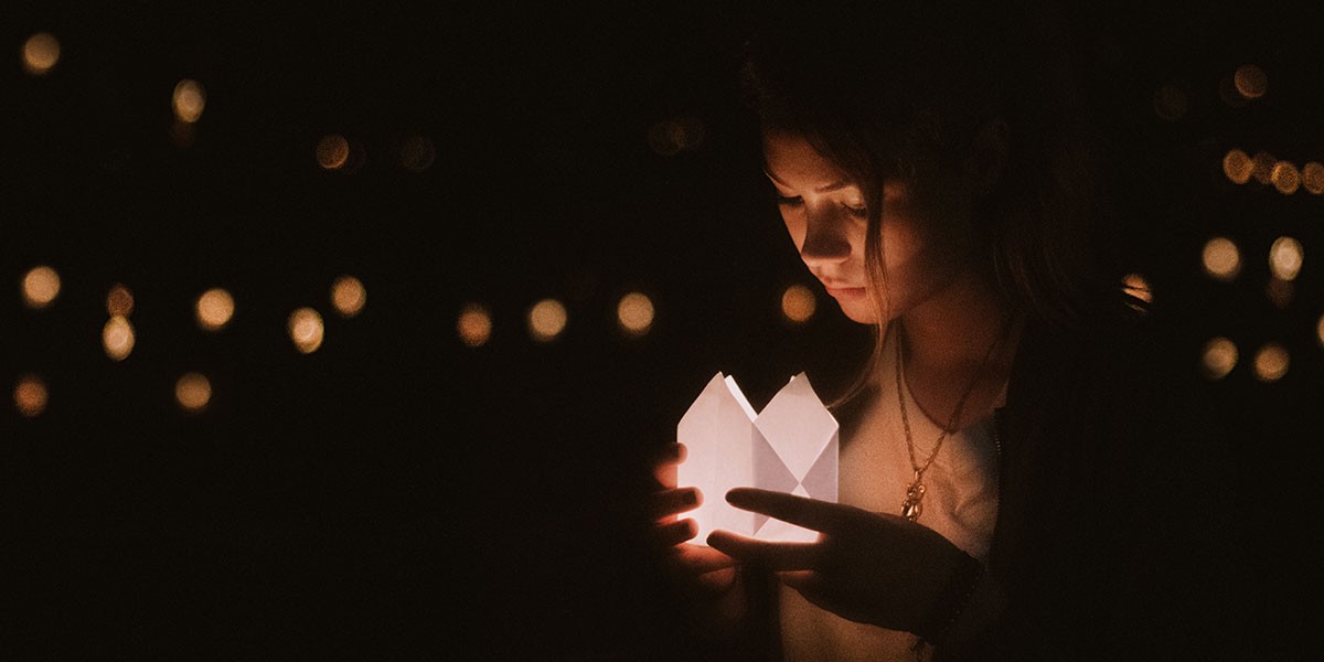 Young woman holding a glowing paper lantern in the dark, with blurred lights in the background.