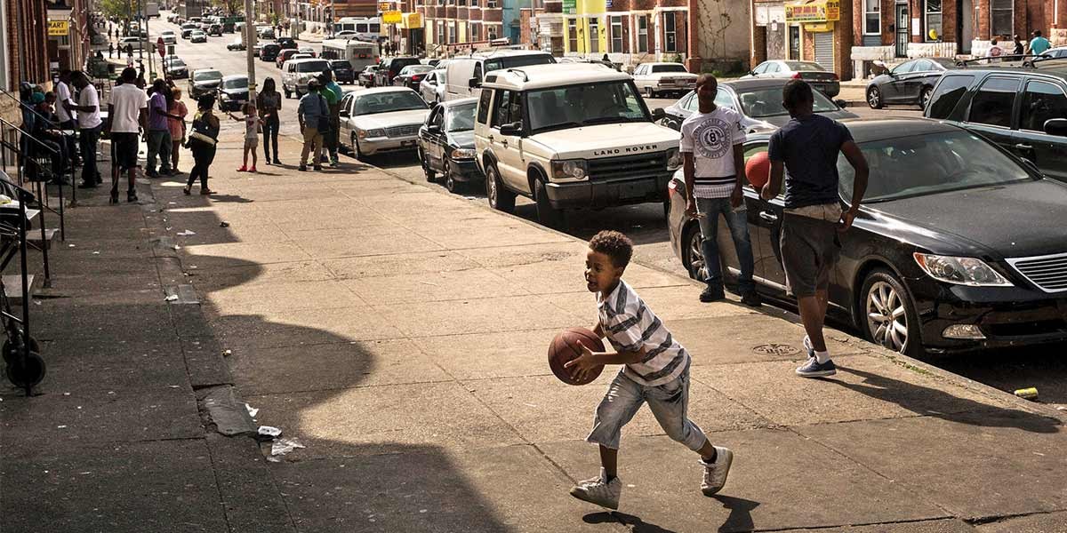 Boy dribbling basketball on a city sidewalk with people and parked cars nearby.