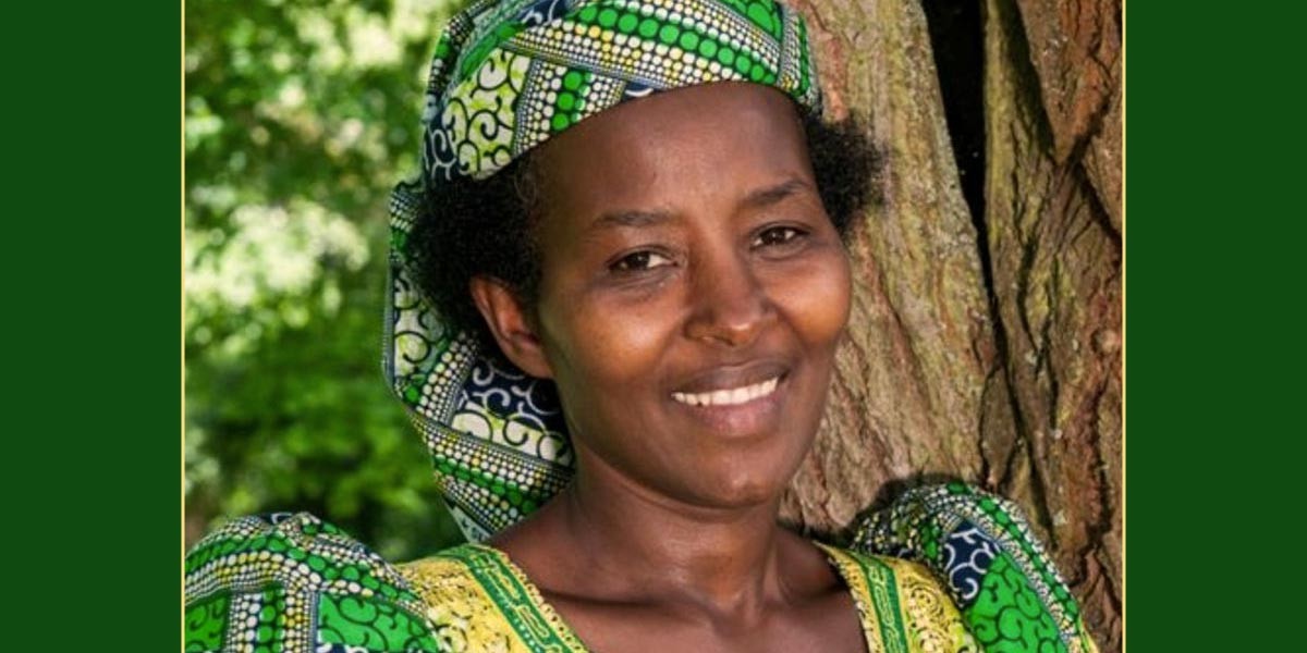 Smiling woman wearing a green and yellow patterned headscarf and dress standing by a tree.