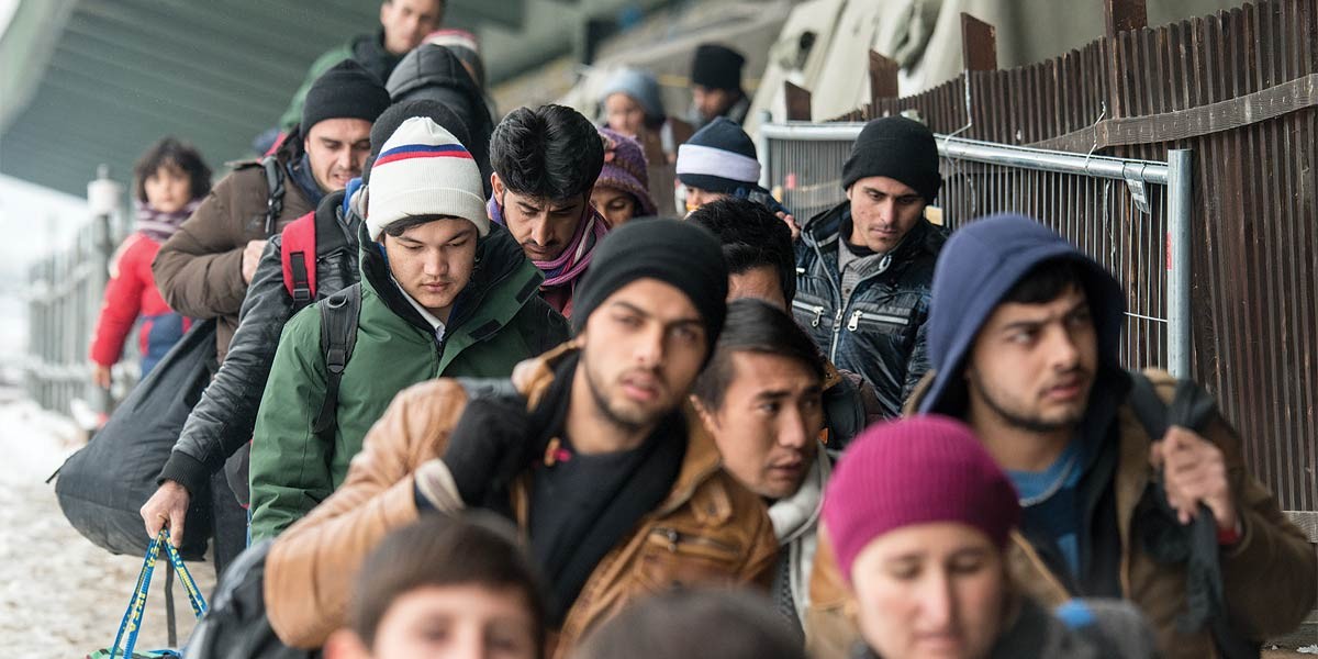 A line of people outdoors wearing winter clothes and carrying bags, standing near a fence.