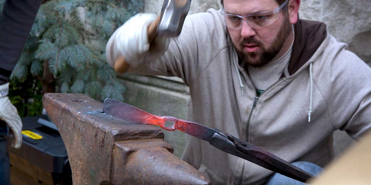 Man wearing safety glasses hammers heated metal on an anvil during blacksmithing.