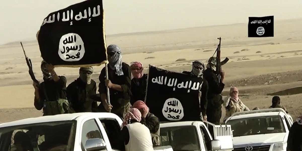 Armed masked men holding black flags with white Arabic script standing on white pickup trucks in a desert.