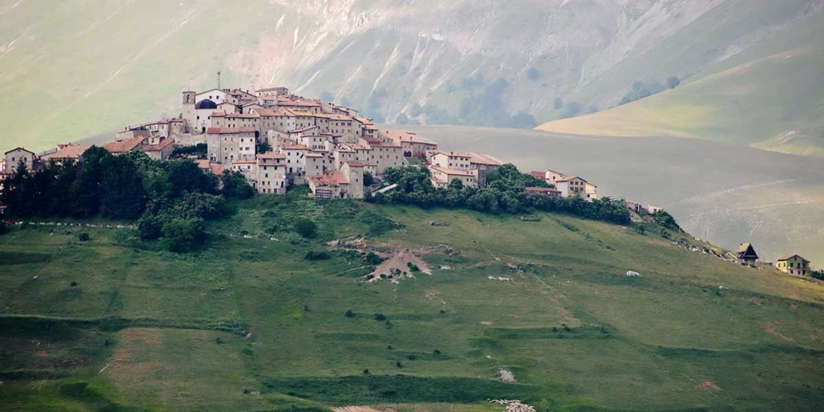 Small village with clustered houses and red roofs atop a green hill surrounded by mountains.