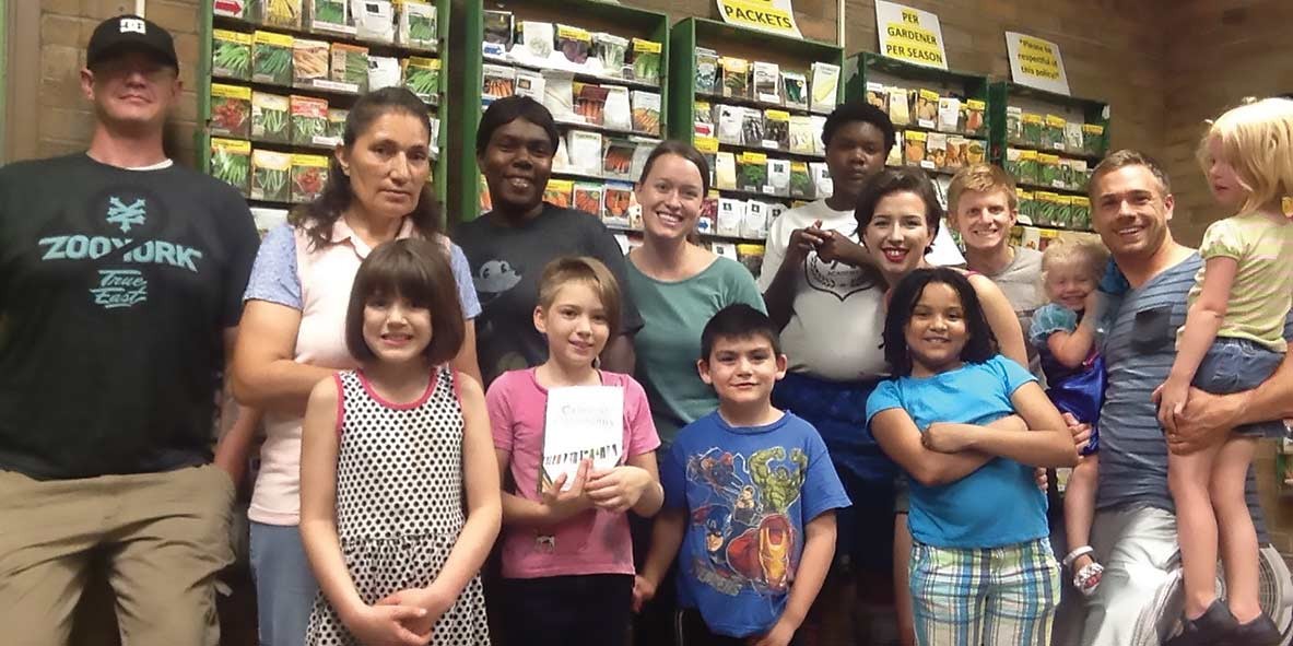 Group of adults and children smiling in front of wall with seed packets in a garden store.
