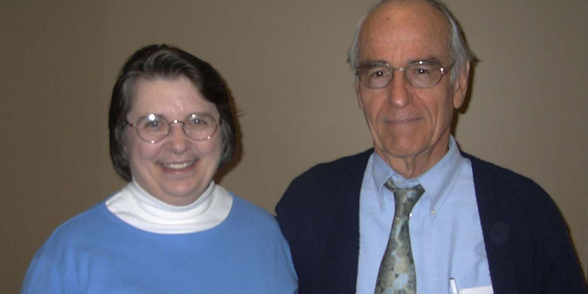 Smiling elderly man and woman standing next to each other against a plain wall.
