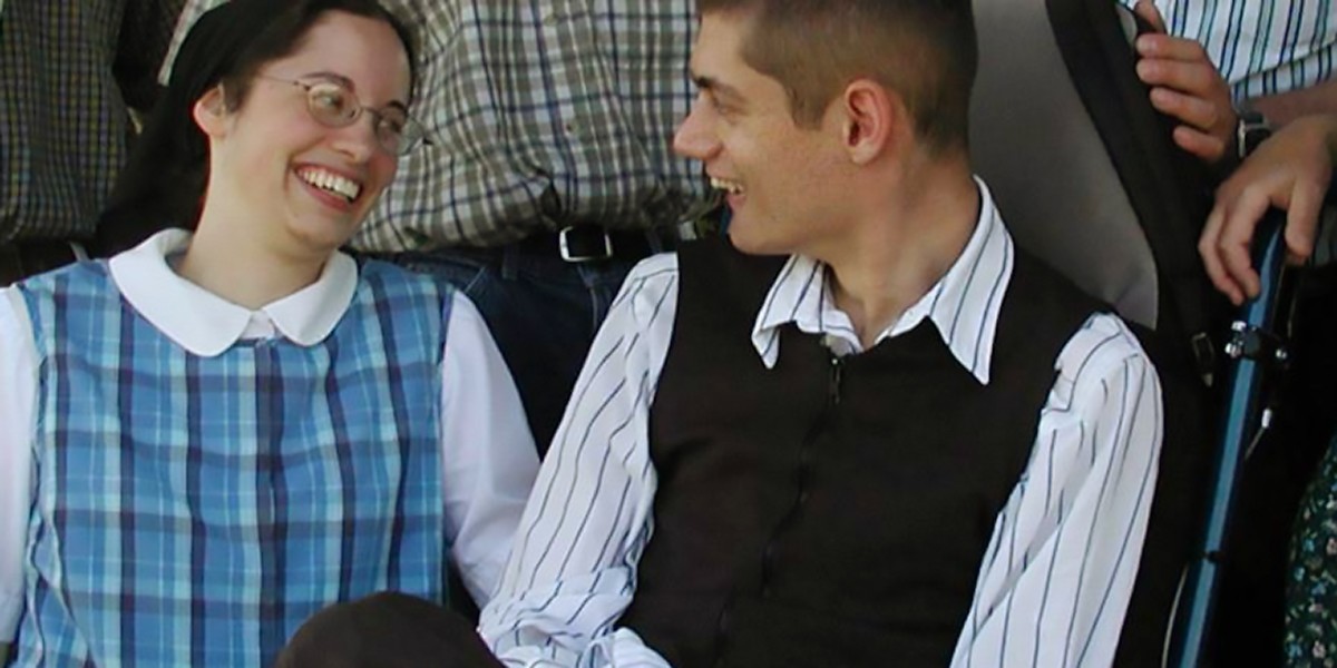 A young woman and man smiling at each other, seated closely, wearing plaid and striped clothes.